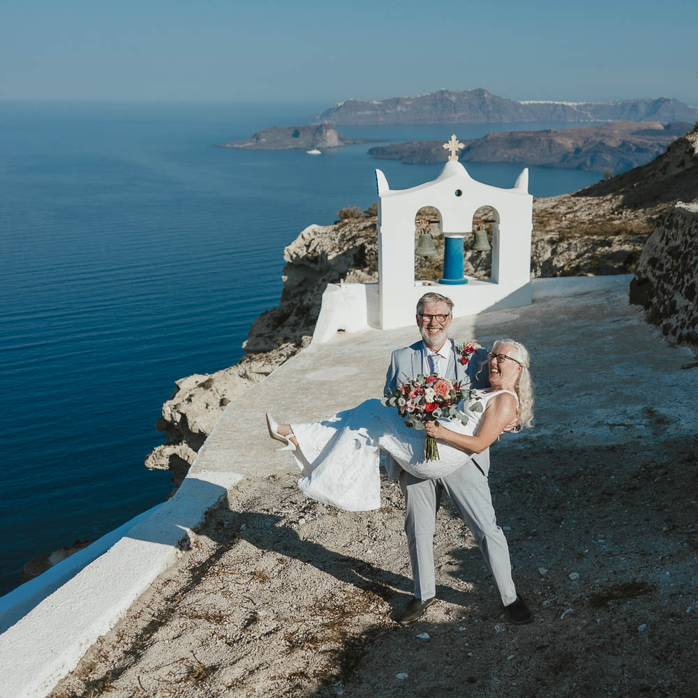 Just like 25 years ago, the husband carries his wife in his arms with the Santorini landscape as background